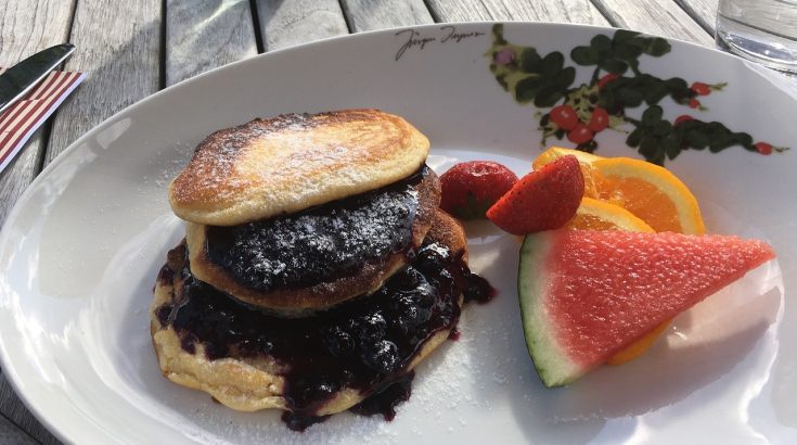 Bialy talerz, na którym znajduje się nalesnik z dżemem a obok kawałki arbuza, pomarańczy i truskawki.