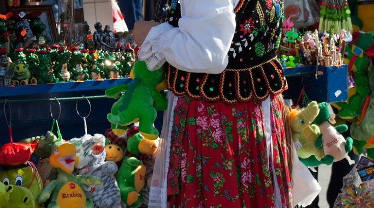 Kobieta w kolorowym, tradycyjnym stroju krakowskim stojąca przed straganem z zabawkami.