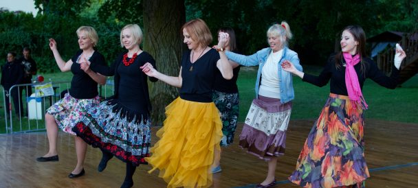 Na obrazku znajduje się grupa kolorowu ubranych kobiet, w różnym wieku, tańczą trzymając się za ręce.