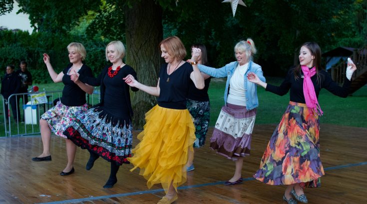 Na obrazku znajduje się grupa kolorowu ubranych kobiet, w różnym wieku, tańczą trzymając się za ręce.
