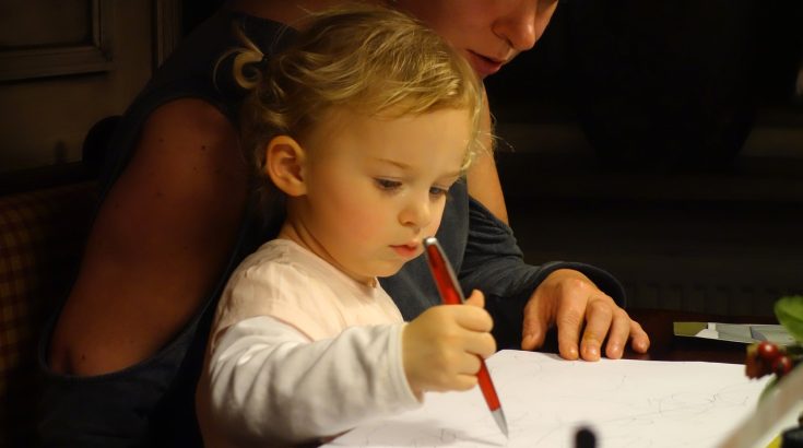 Obrazek przedstawia matkę siedzącą przy biurku z dzieckiem. Dziecko trzyma czerwony długopis w prawej ręce.