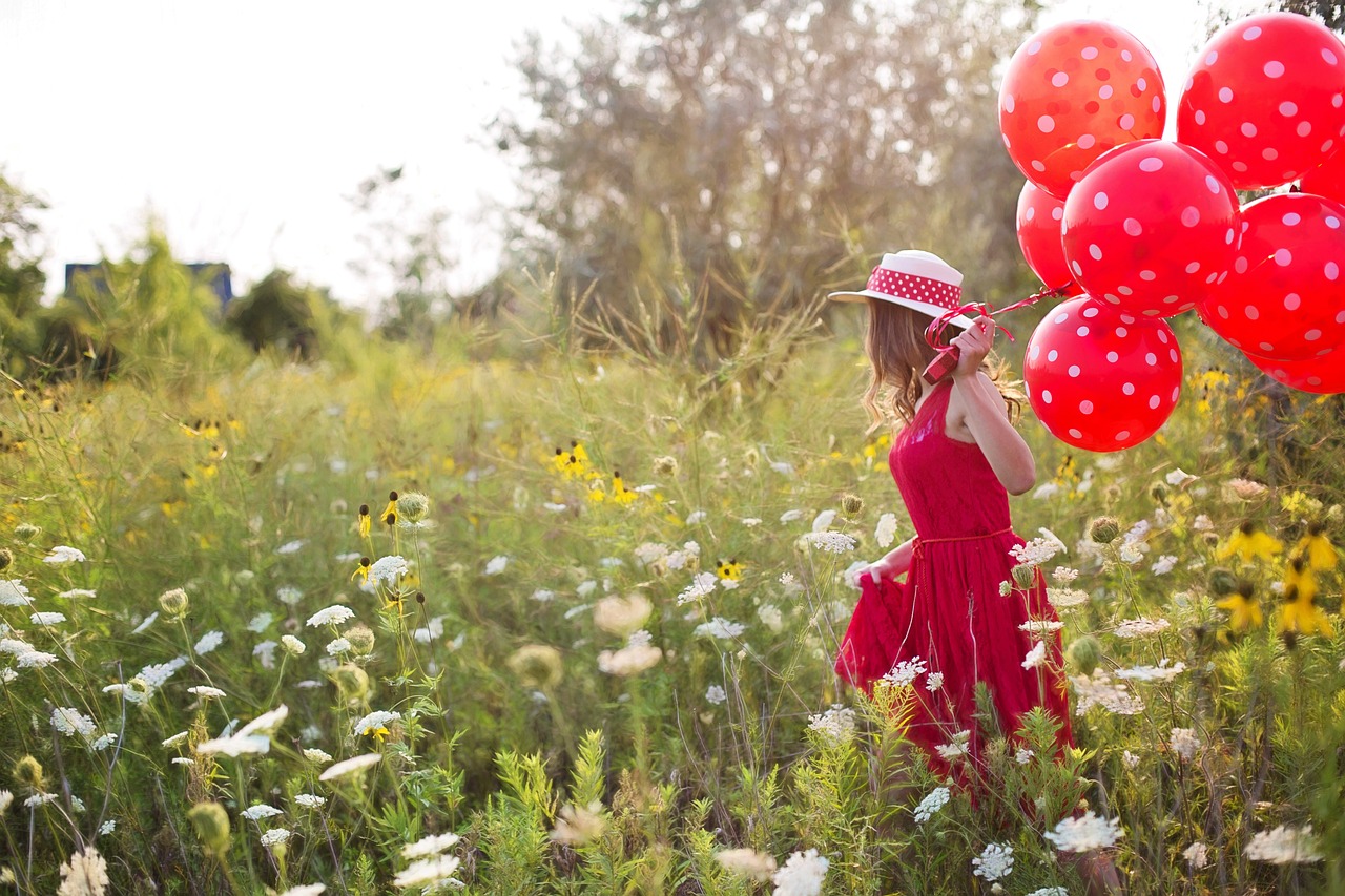 Na obrazku znajduje się dziewczynka w kapeluszu i kolorowej sukience, trzyma w ręce czerwone balony, idzie po łące.