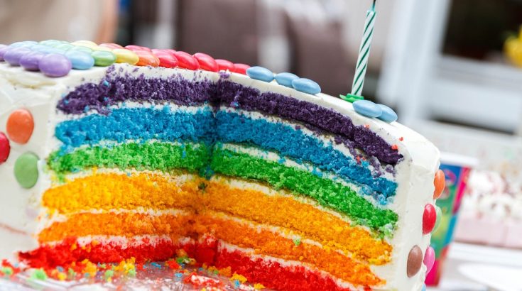 Zdjęcie przedstawia tort z kolorowymi warstwami i świeczką.